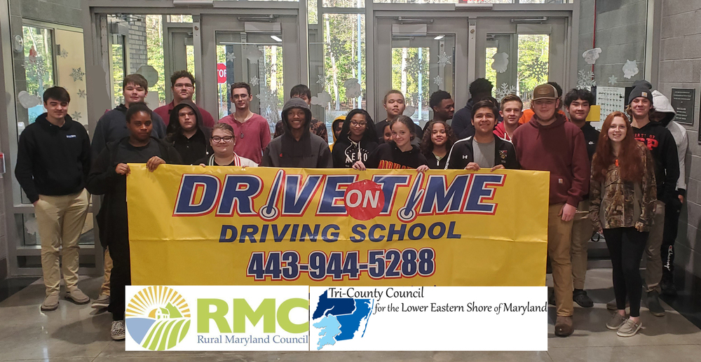 A group of 20+ teenage students standing in the lobby of the Tech School holding a banner with text that reads: Drive On Time Driving School 443-944-5288 and the Rural MD Council and Tri County Council of the LES logos