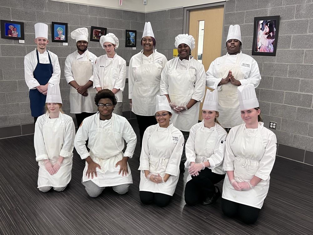 A group of students in chef hats and white aprons separated into two rows in the technical high school banquet room