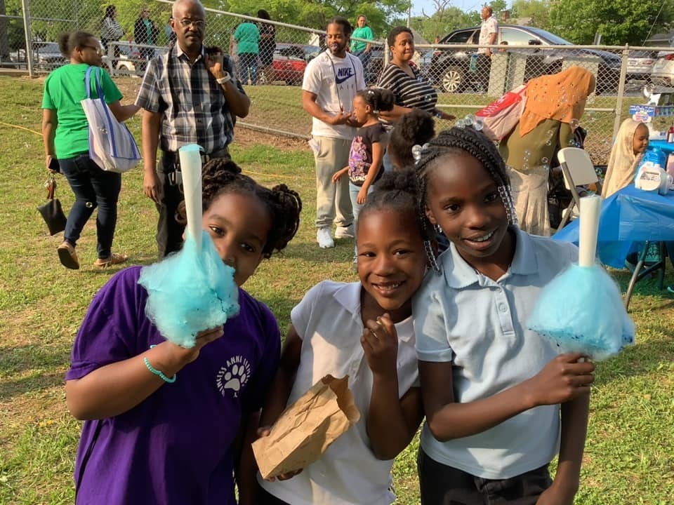 three dark skinned girls two of which are holding cotton candy and smiling, behind them is a playground with lots of people on it
