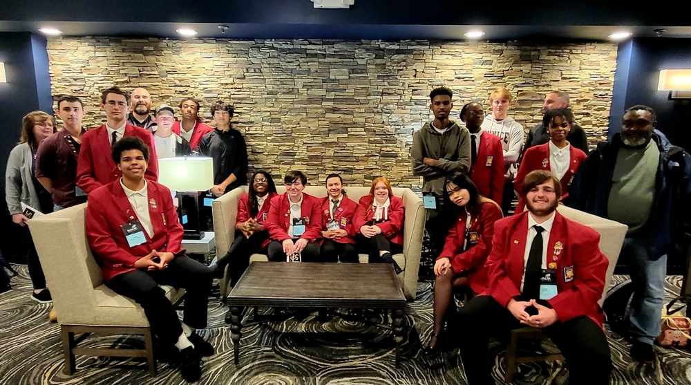 a group of students in red sports coats with emblems that read SkillsUSA sitting on various couches with groups of adults standing behind them against a stone background
