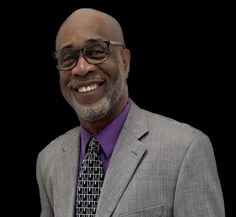 dark skinned man with beard and glasses in a gray suit, smiling
